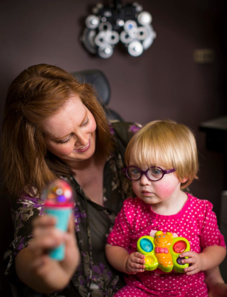 Woman is Holding a Toy in Front of a Child Wearing Glasses
