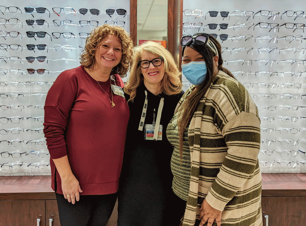 three woman stand in front of a wall of eyeglasses