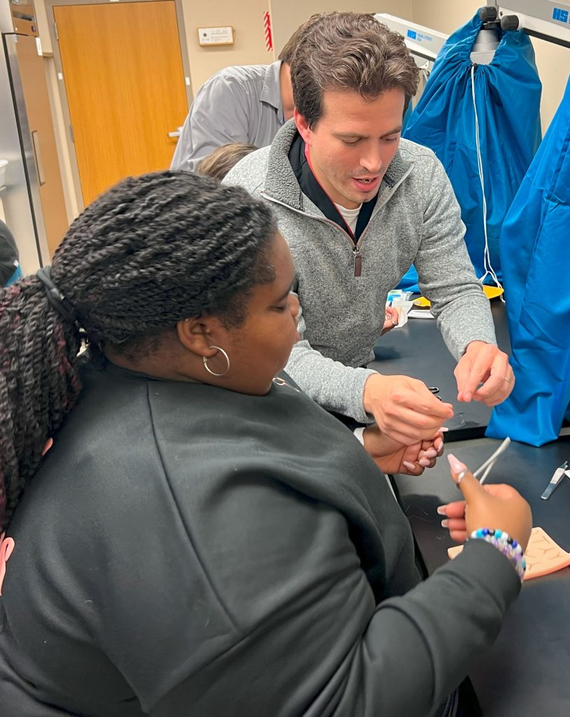 a man in a grey sweatshirt shows a woman in a black jacket how to suture