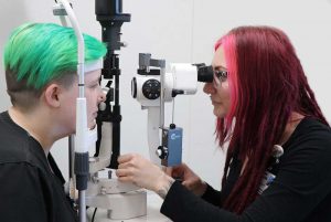 a woman with long pink hair examines a woman's eyes through a machine
