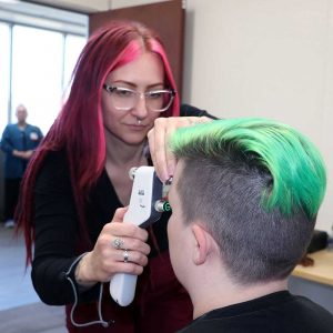 a woman with long pink hair examines a woman's eyes