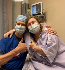 a man in blue surgical scrubs and a woman in a purple hospital gown give 'thumbs up'