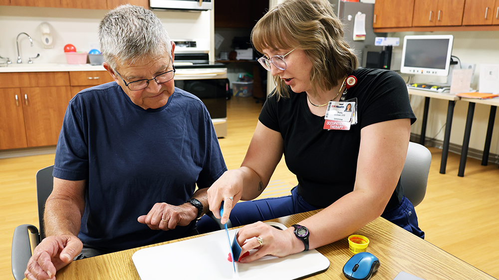a woman in a black shirt demonstrates safe cutting practices while a man in a blue shirt looks on